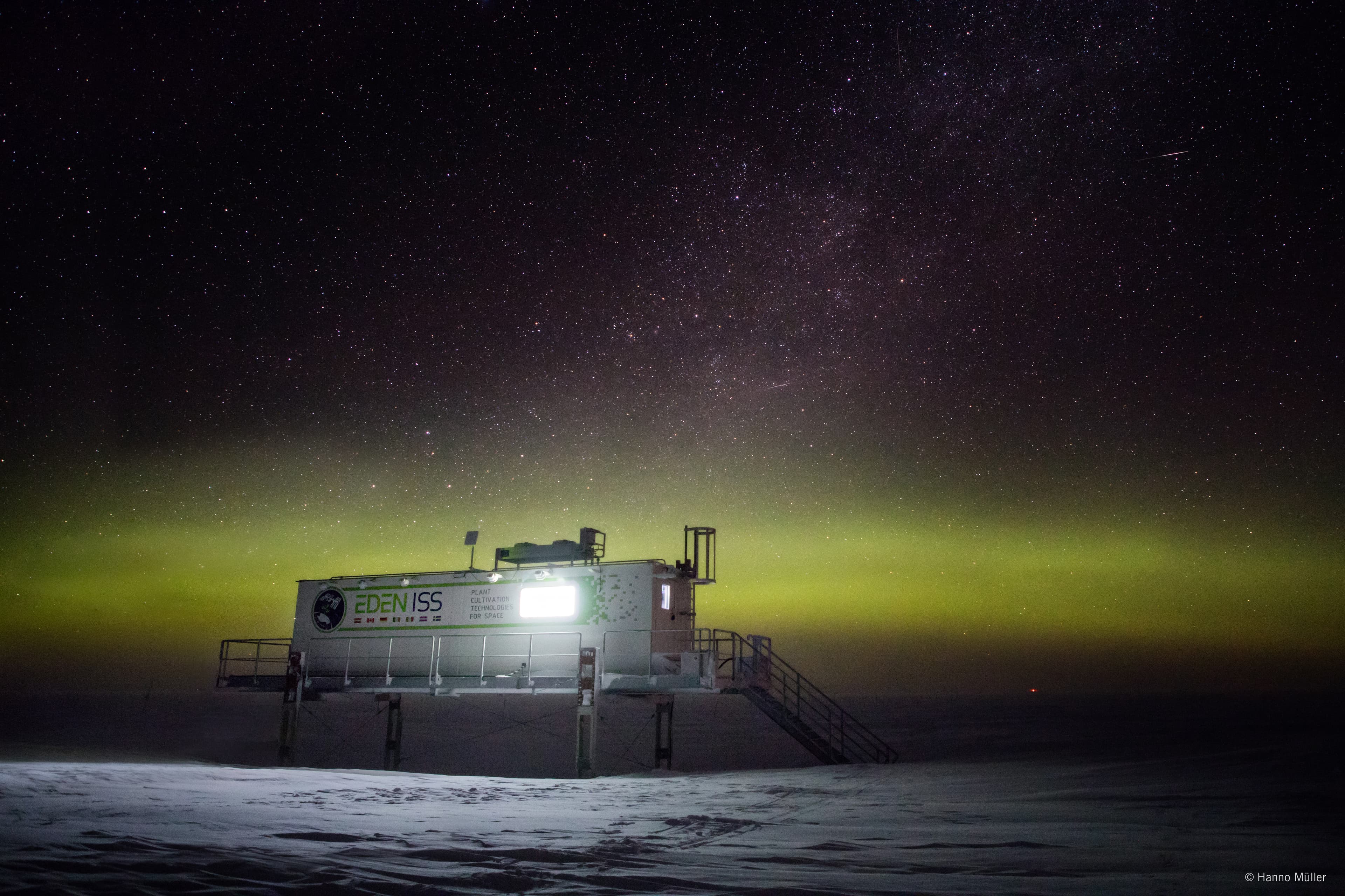 Snowy landscape with a raised research module labeled 'EDEN ISS', lit windows and a stairway, against a green aurora and a star-filled night sky.