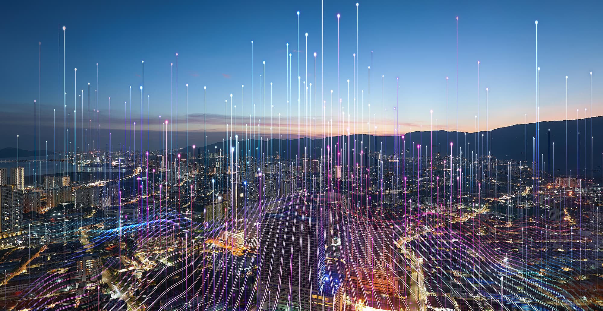Night cityscape with glowing skyline and roads, overlaid by vertical, colorful light streaks stretching upward toward a twilight sky with distant mountains.